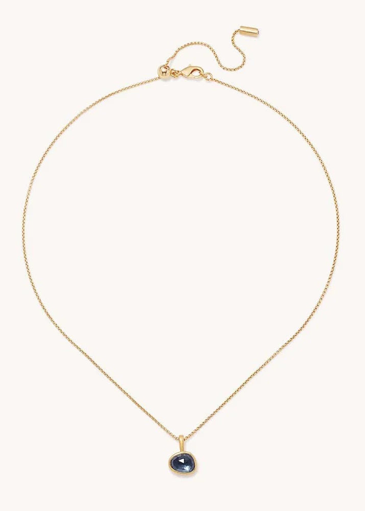 A delicate gold chain necklace with a small teardrop-shaped dark blue gemstone pendant, displayed on a clean off-white background.