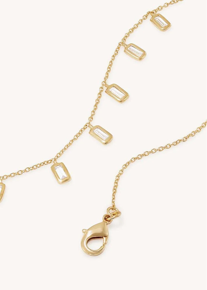 A delicate gold chain necklace featuring multiple small rectangular bezel-set clear stones along the chain, with a lobster clasp visible at one end, photographed on a clean off-white background.