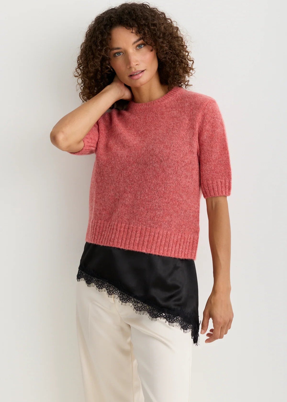 A woman modeling a short-sleeve knit sweater in heathered dusty rose pink, styled with a black satin underlayer and cream trousers against a plain light background.