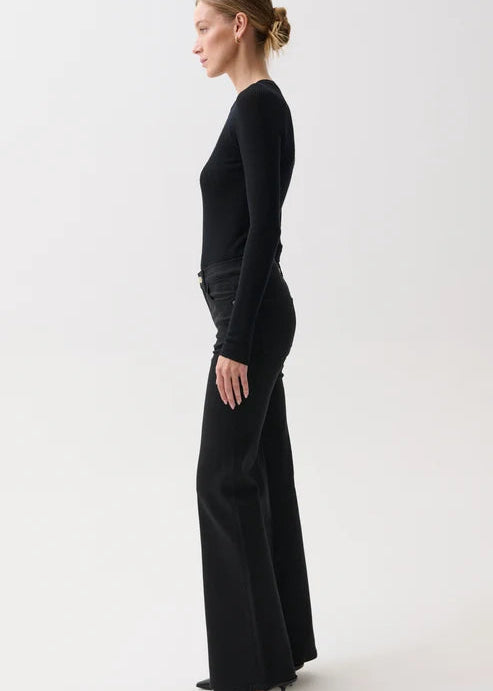 A woman in a side profile wearing a black long-sleeve top and matching wide-leg trousers against a plain light gray studio background.