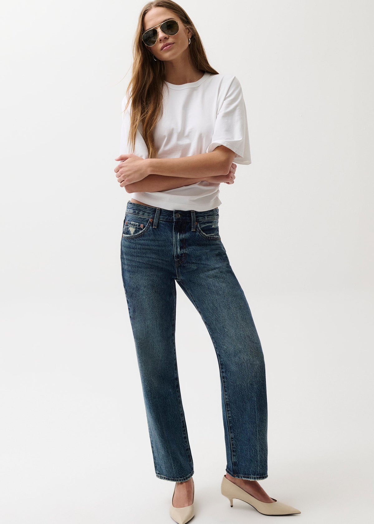 A woman modeling a pair of dark blue straight-leg jeans with a white t-shirt and beige heels, standing against a solid white background.