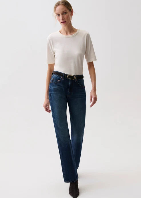 A woman modeling a pair of dark blue straight-leg jeans with a black belt, paired with a cream short-sleeve t-shirt and black ankle boots, against a plain white studio background.