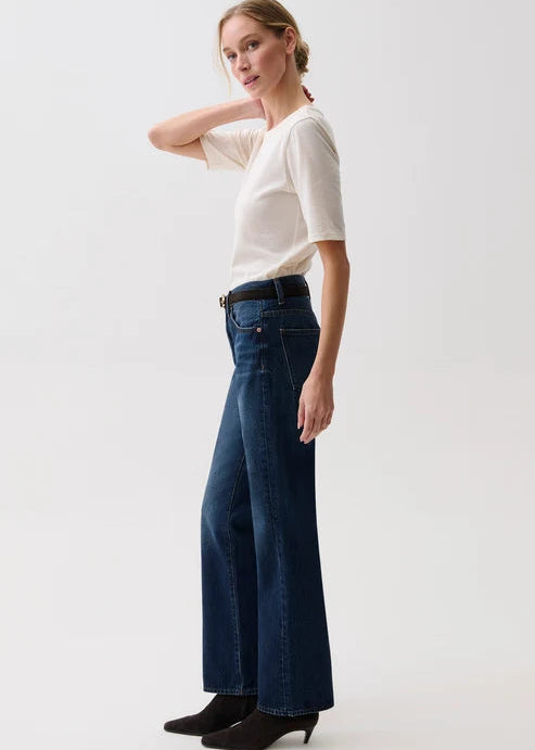 A woman modeling a pair of dark blue wide-leg jeans with a black belt, paired with a cream short-sleeve top and black ankle boots, against a plain light gray studio background.