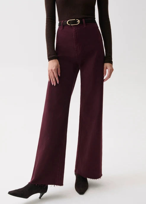 A model wearing high-waisted, wide-leg trousers in a deep burgundy color, styled with a black belt, a dark brown long-sleeve top, and black pointed-toe ankle boots against a light gray studio background.