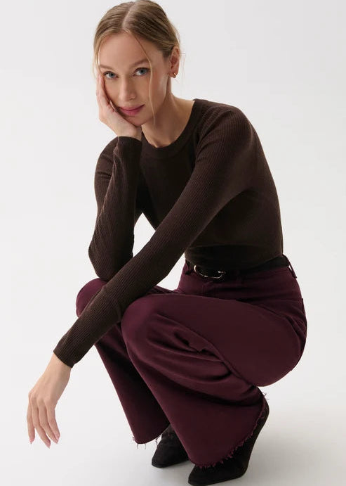 A woman crouching in a studio setting, wearing a dark brown long-sleeve top and deep burgundy wide-leg pants with black shoes.