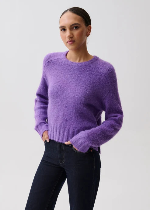 A woman modeling a fuzzy, short-sleeve crewneck sweater in a soft lavender-purple color, paired with dark blue jeans, against a plain light gray studio background.