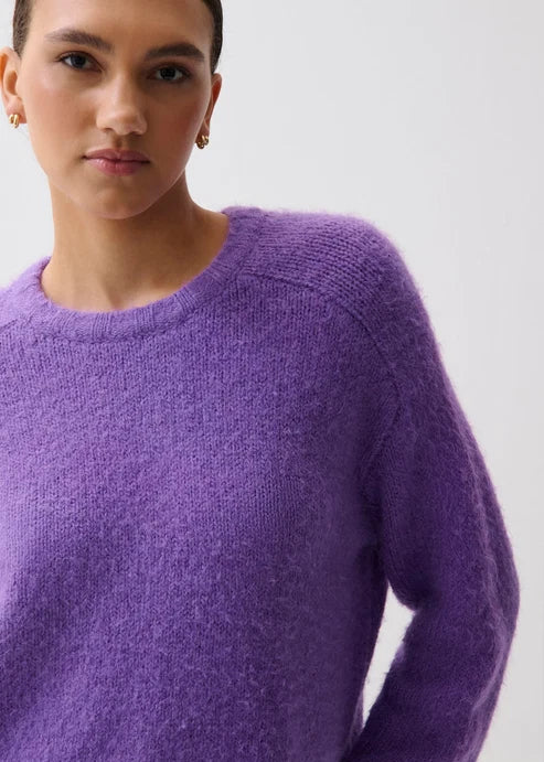 A close-up of a model wearing a fuzzy, textured purple crewneck sweater against a plain light gray background.