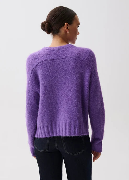A woman shown from the back wearing a fuzzy purple knit sweater and dark jeans against a plain light gray studio background.