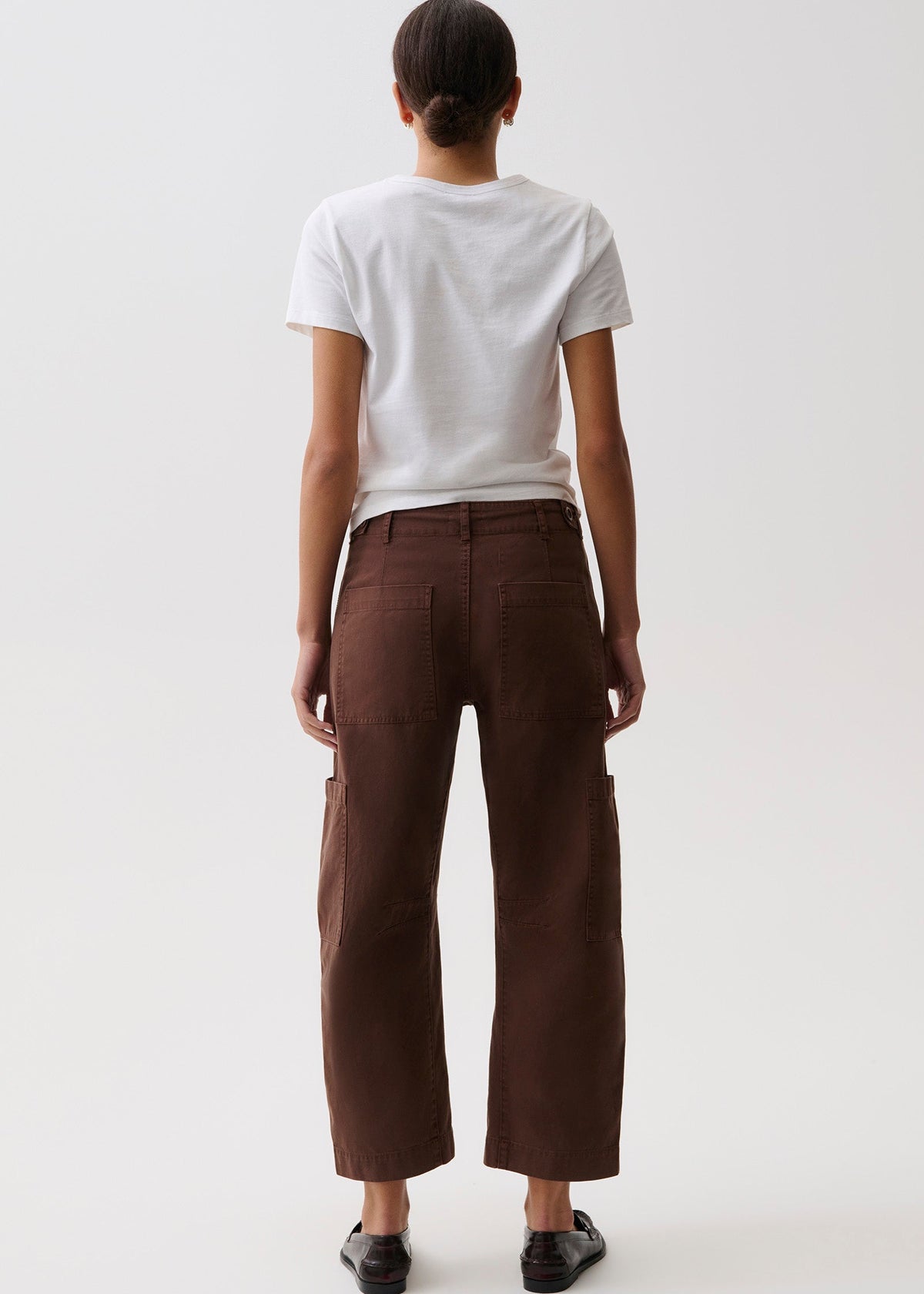 A model shown from the back wearing dark brown cropped cargo pants, a white t-shirt, and black shoes against a plain white studio background.
