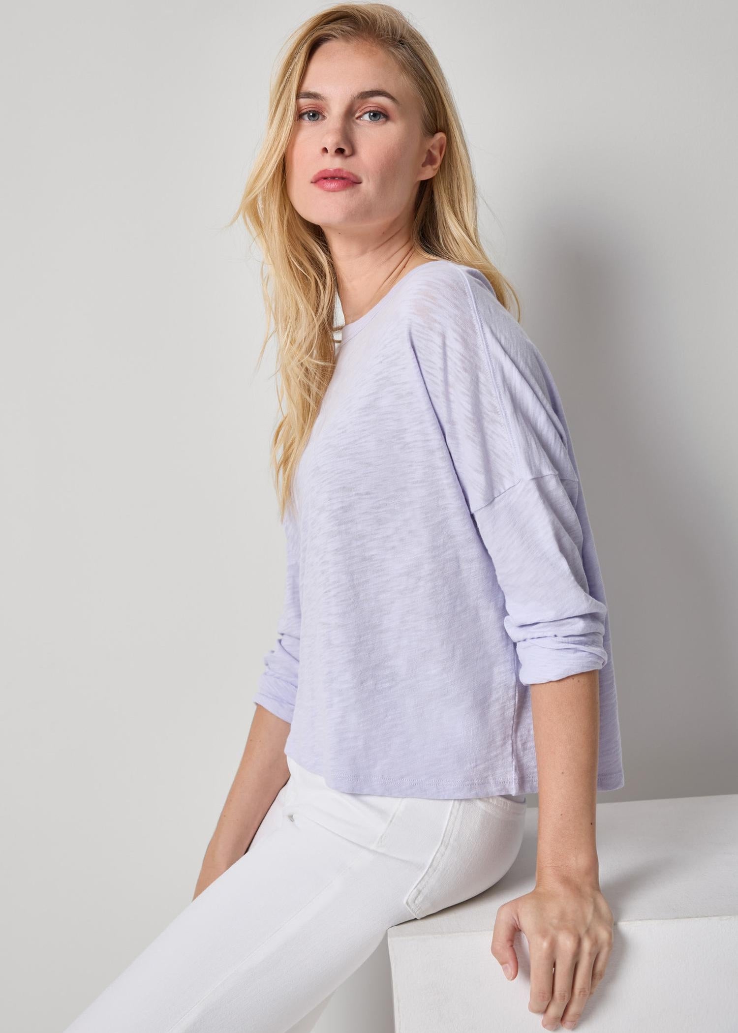 Woman wearing a light purple sweater and white pants against a plain background