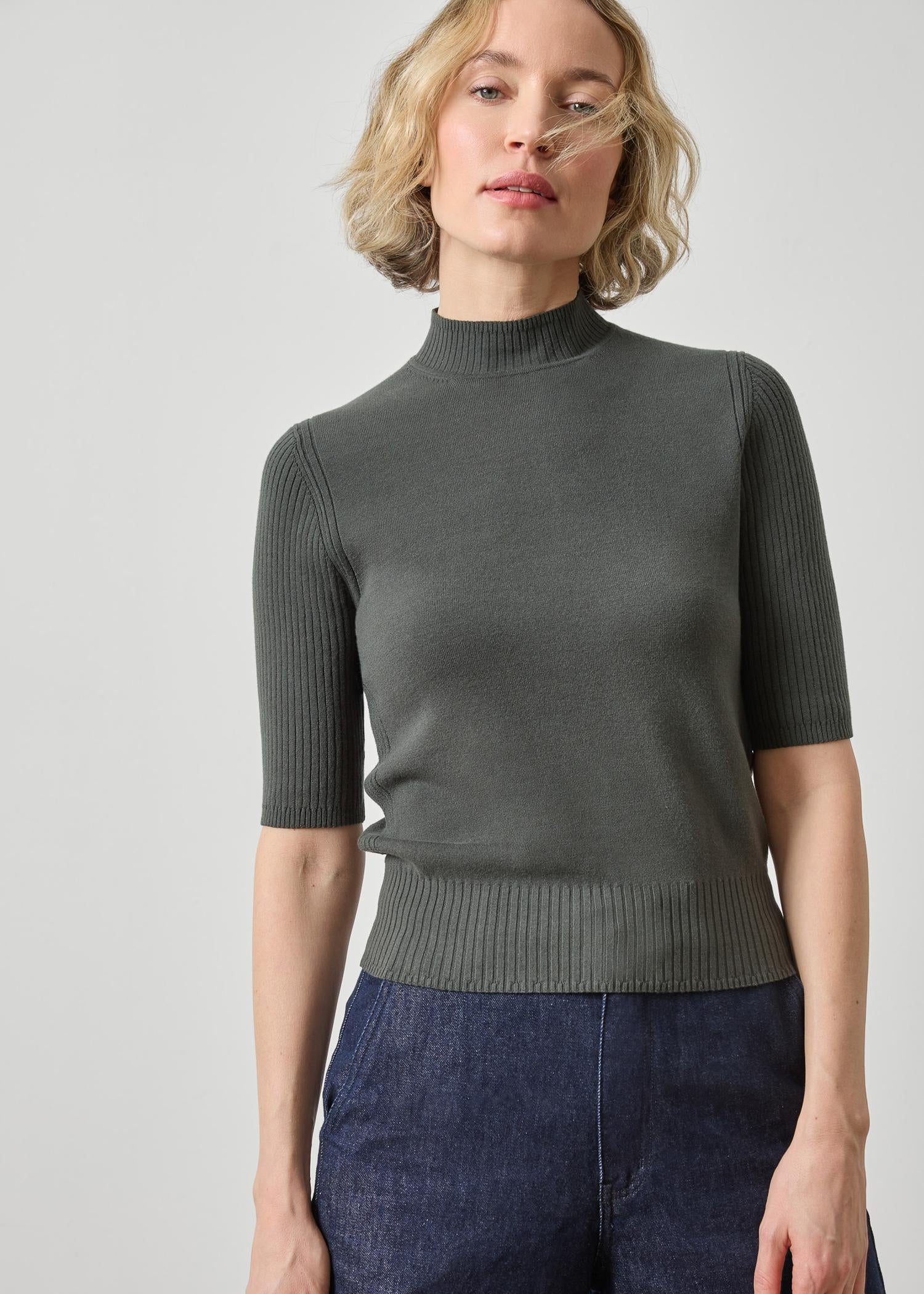 A woman wearing a dark gray ribbed short-sleeve mock neck sweater and dark blue jeans, photographed against a light gray studio background.