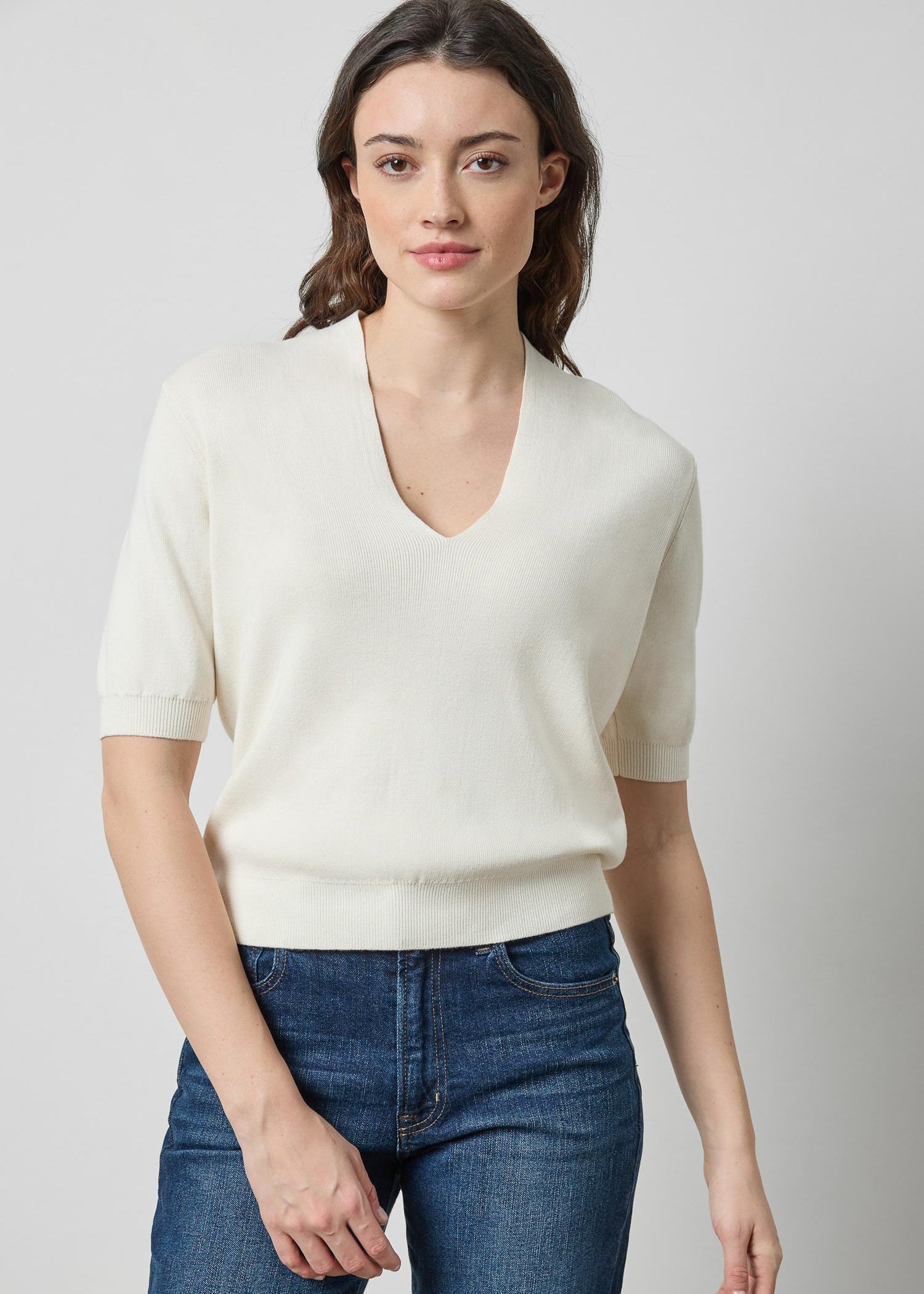 A woman modeling a short-sleeve cream knit V-neck sweater with blue jeans, photographed against a plain light gray studio background.