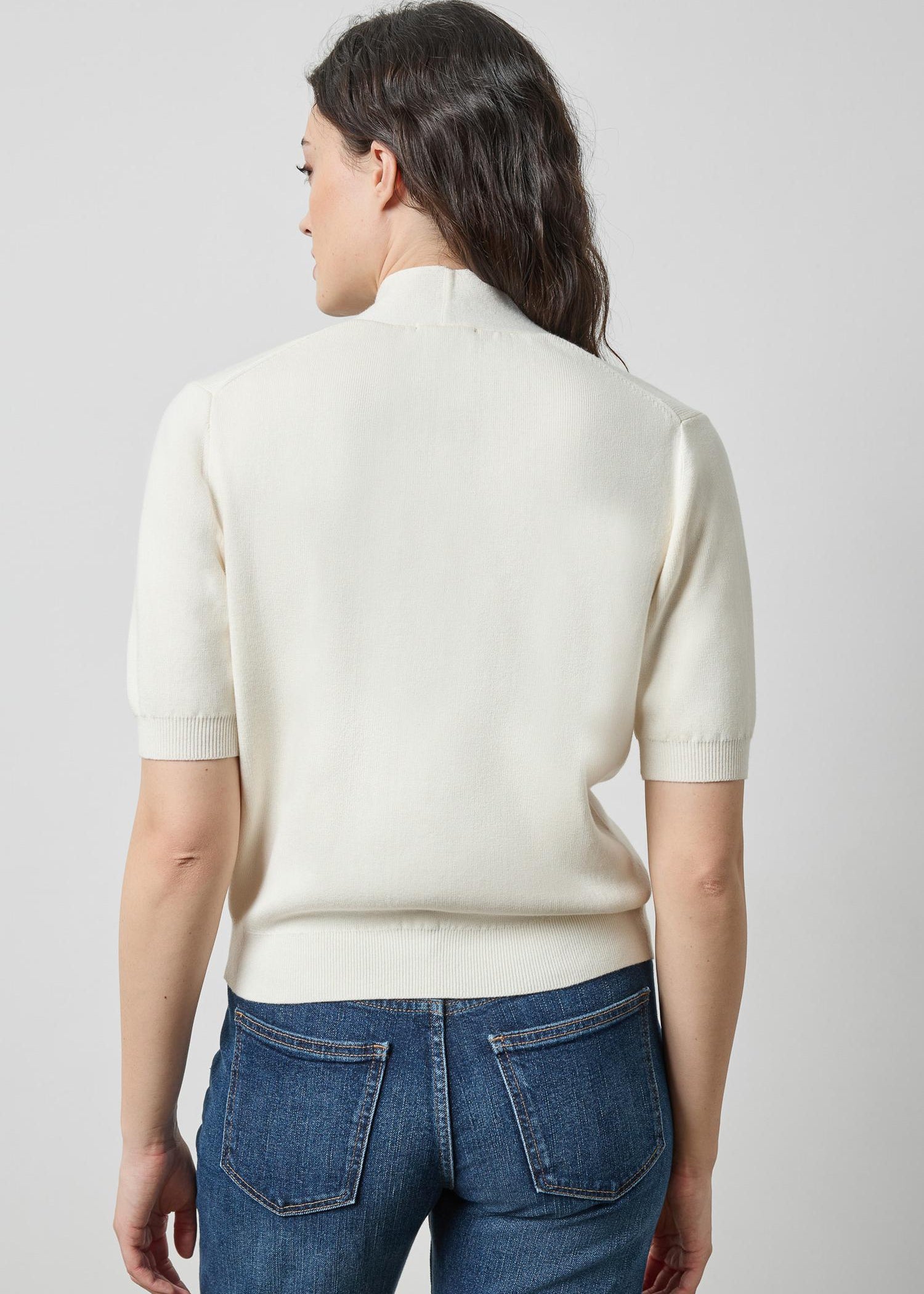 A woman viewed from the back wearing a cream-colored short-sleeve knit mock neck sweater and blue jeans against a light gray studio background.