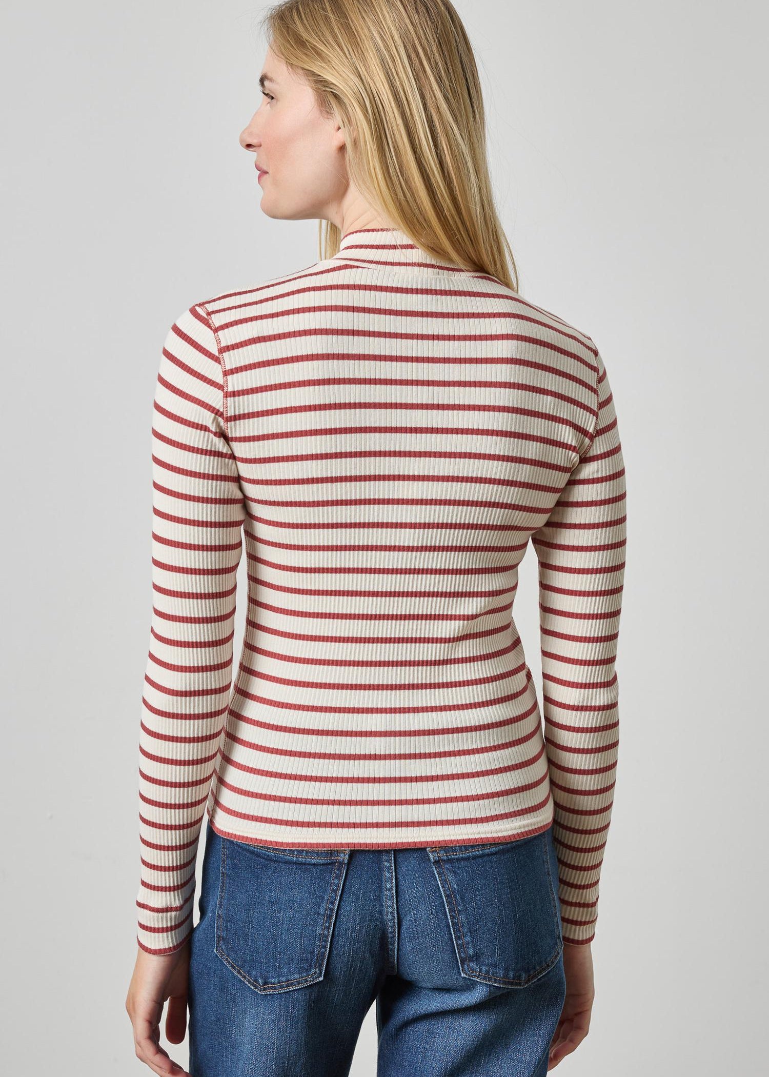 A woman shown from the back wearing a fitted long-sleeve ribbed top with thin horizontal red stripes on a warm beige or cream base, paired with blue jeans, against a light gray studio background.