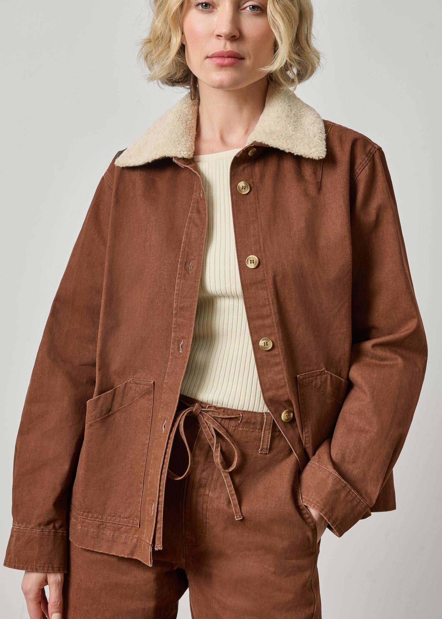 A model wearing a brown corduroy jacket with a cream sherpa collar, paired with matching brown pants and a cream ribbed top, shot against a neutral gray background.
