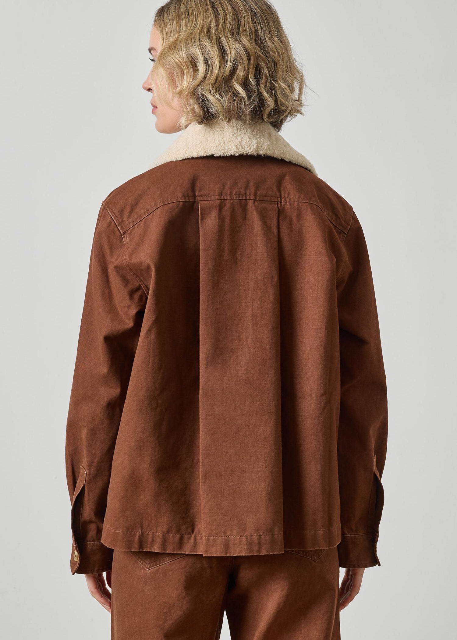 A model shown from the back wearing a brown utility-style jacket with a cream-colored shearling collar, paired with matching brown trousers, against a plain light gray studio background.