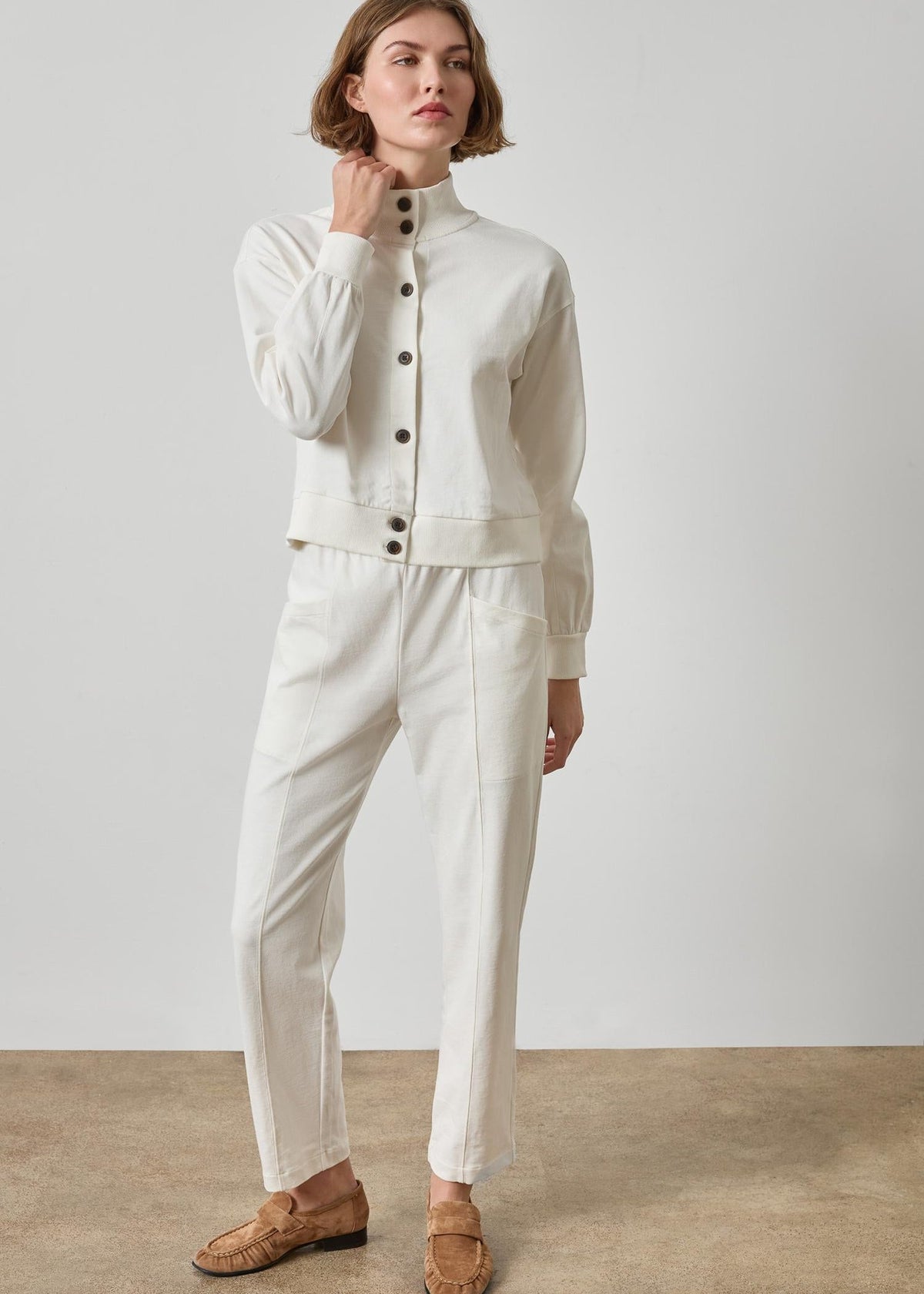 A model wearing a matching off-white two-piece set consisting of a cropped button-front cardigan and straight-leg trousers, paired with brown woven loafers, against a neutral studio backdrop.