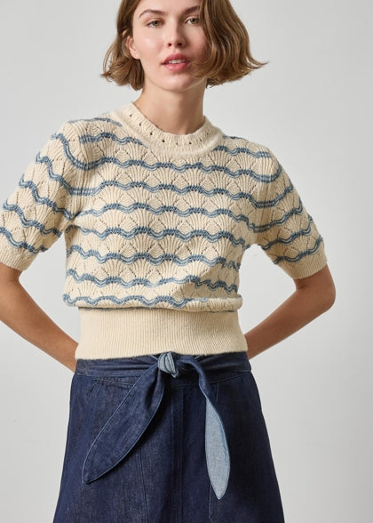 A woman wearing a cream short-sleeve knit sweater with blue wavy stripe patterns and a matching ribbed hem, paired with a dark blue denim skirt, against a light gray studio background.