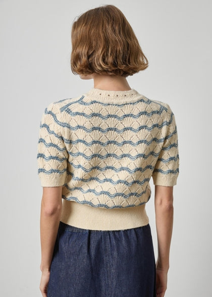 A woman shown from the back wearing a cream knit short-sleeve top with a blue wavy stripe pattern and a matching ribbed hem, paired with a dark blue skirt, against a light gray studio background.