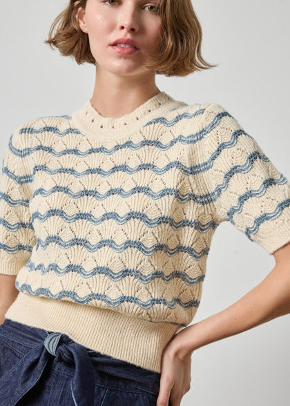 A woman wearing a cream-colored short-sleeve knit sweater with blue wavy horizontal stripes, paired with dark blue jeans, against a light gray studio background.