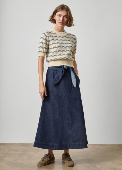 A model stands against a plain light gray studio wall wearing a cream knit short-sleeve top with blue-gray wavy stripes and a dark blue midi skirt with a tie waist, paired with brown shoes.