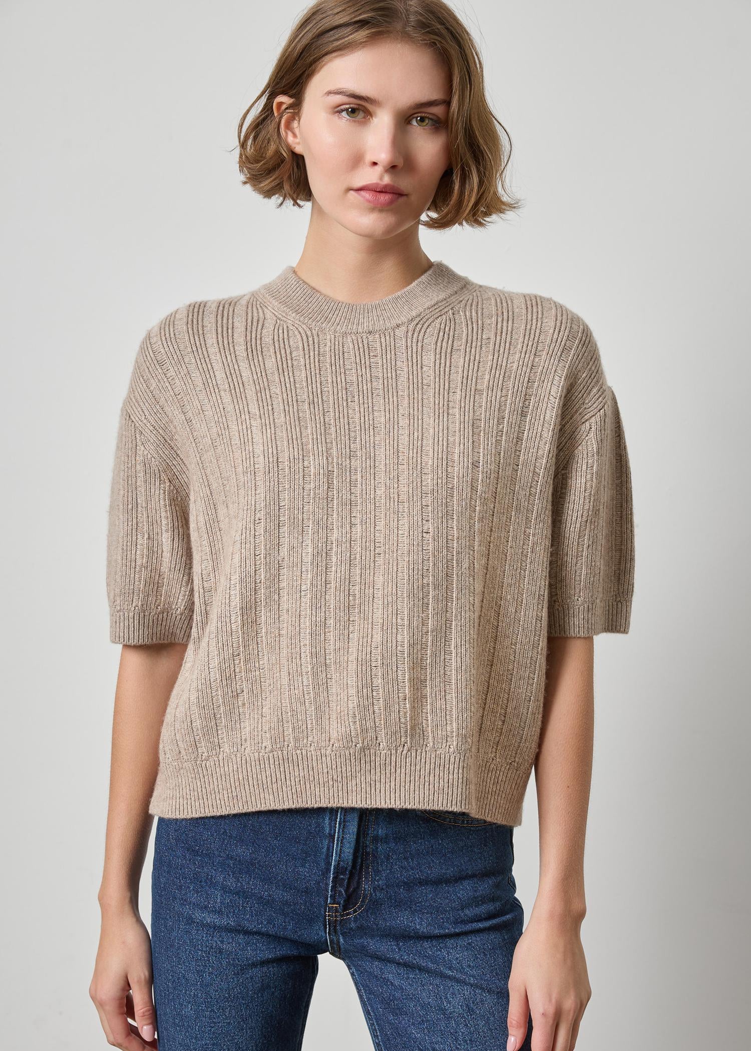 Woman wearing a beige ribbed sweater and blue jeans on a plain background