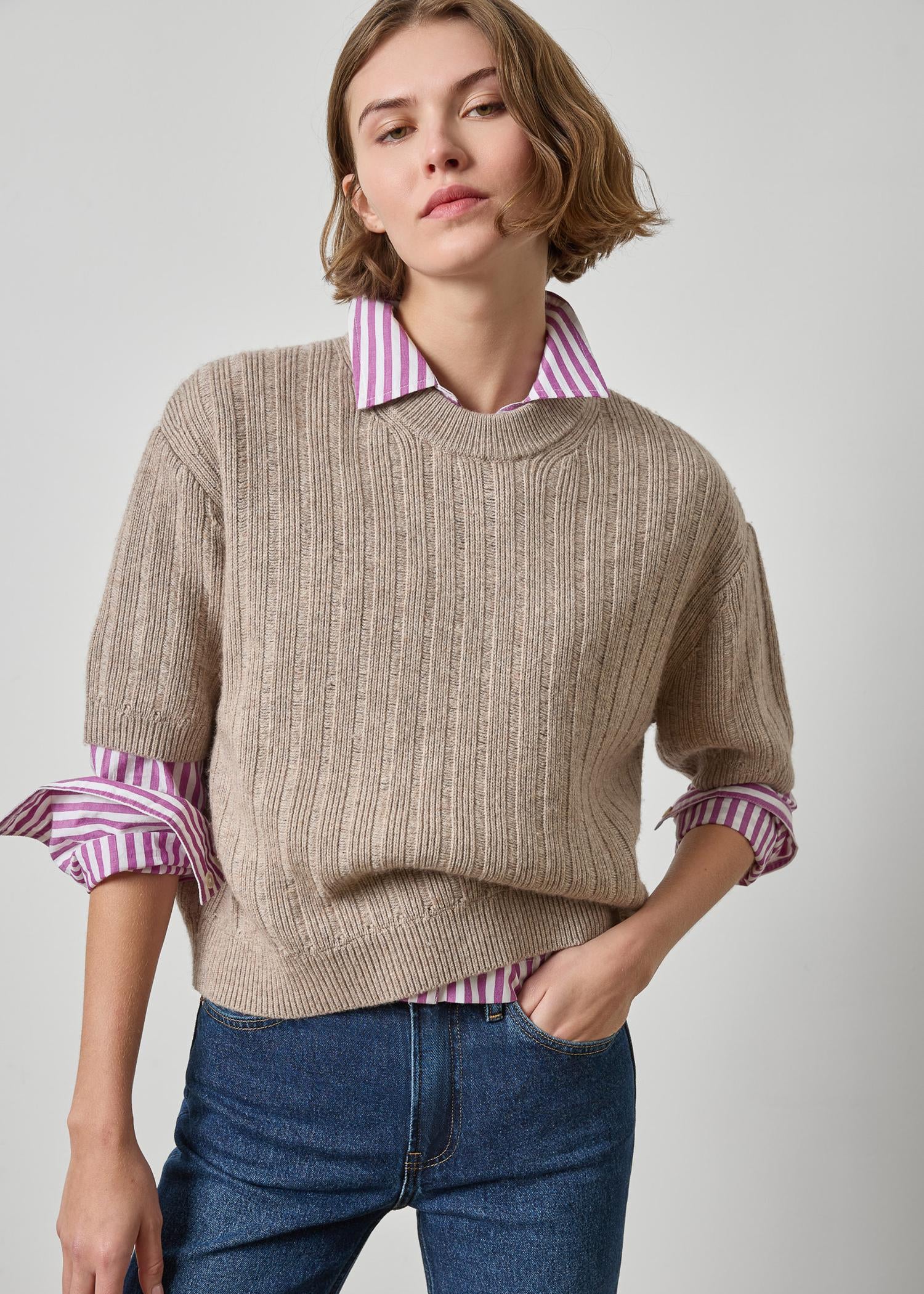 Woman wearing a beige sweater with striped shirt collar and blue jeans on a light gray background