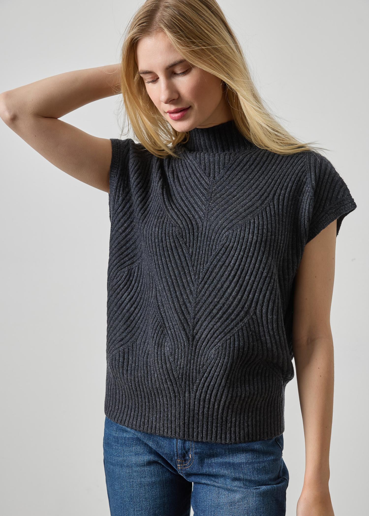 A woman wearing a dark charcoal gray ribbed knit sleeveless mock neck sweater vest with blue jeans, posing against a light gray studio background.