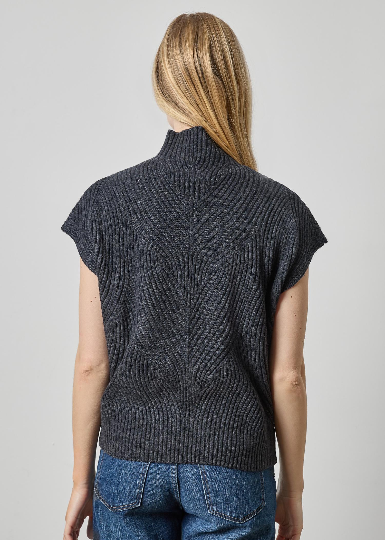 A woman viewed from the back wearing a dark charcoal gray ribbed knit mock-neck sweater with short cap sleeves and blue jeans, against a light gray studio background.