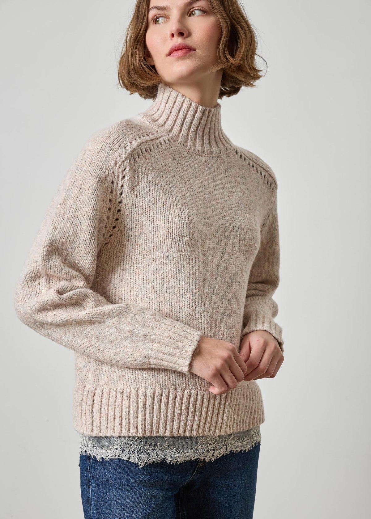 A woman wearing a beige ribbed turtleneck sweater with textured knit details on the shoulders, paired with blue jeans, against a light gray studio background.