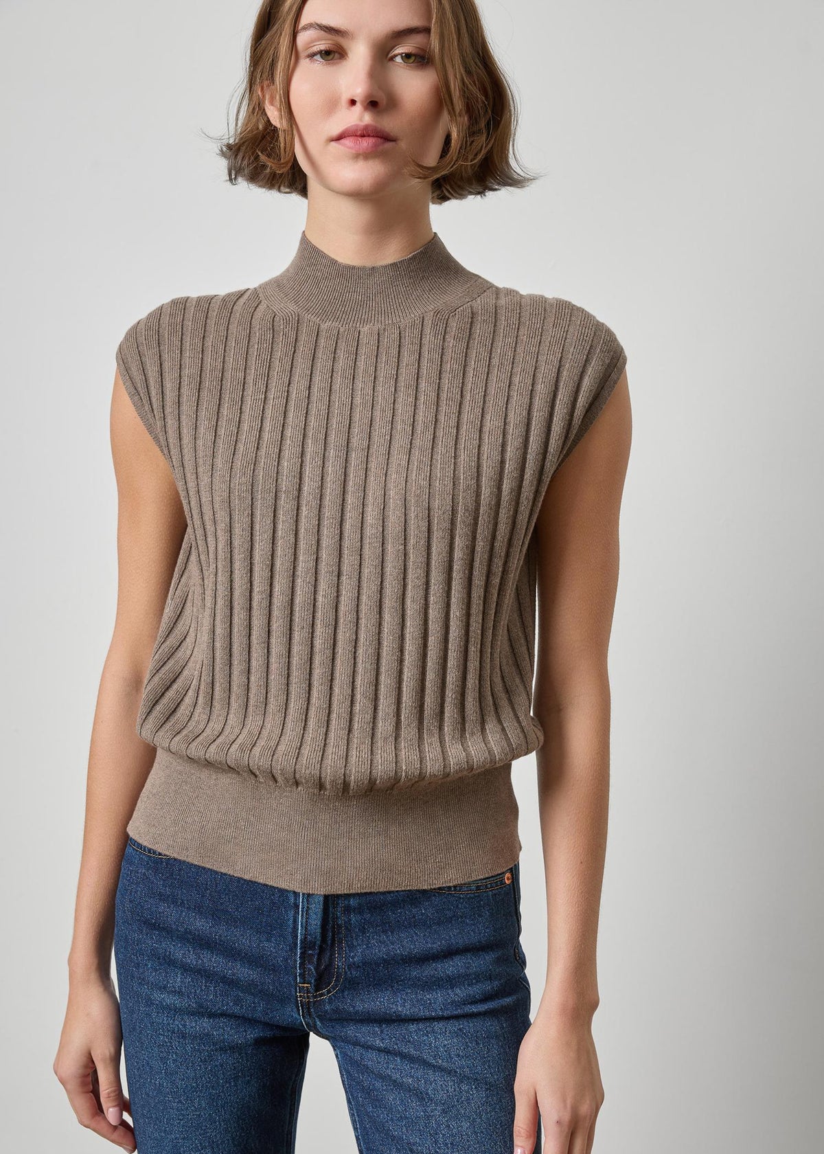 A woman wearing a taupe ribbed knit mock neck sleeveless sweater vest paired with blue jeans, photographed against a neutral light gray studio background.