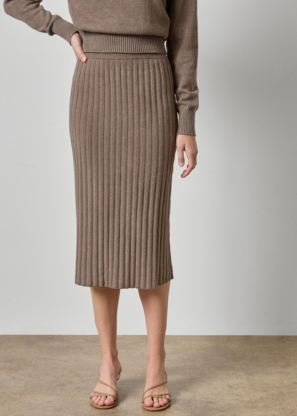 A model wearing a taupe brown ribbed knit midi skirt and matching knit top, paired with beige strappy heels, against a neutral studio backdrop.