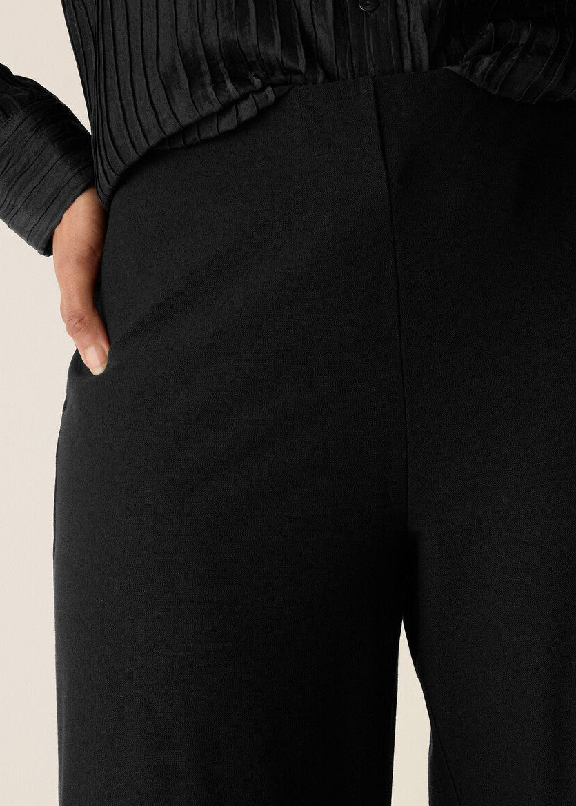 Close-up of a person wearing black wide-leg trousers with a hand in the pocket, paired with a black pleated top, against a light beige background.