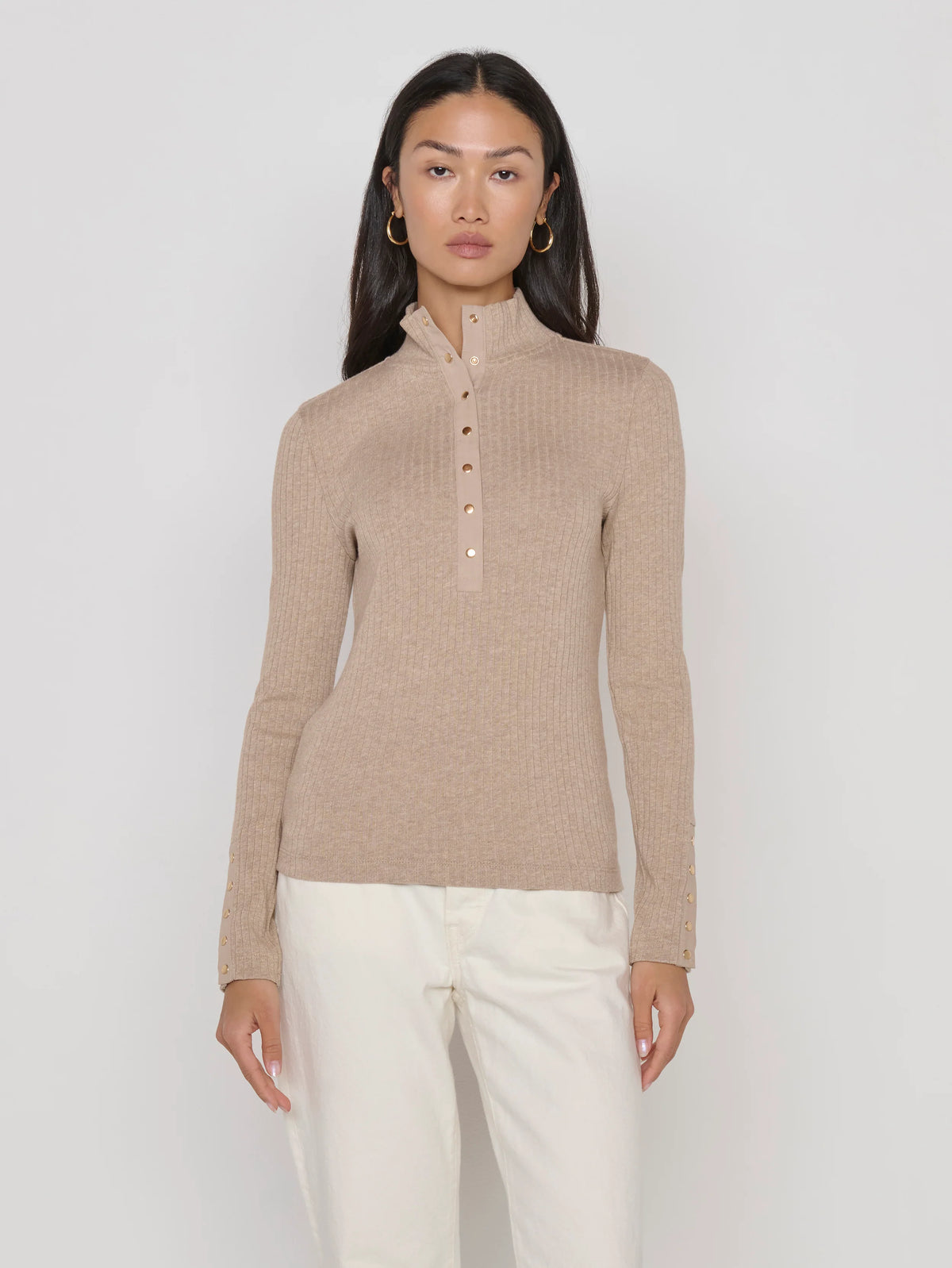 A woman modeling a beige ribbed mock-neck long-sleeve top with a button placket, paired with white pants, against a plain light gray studio background.