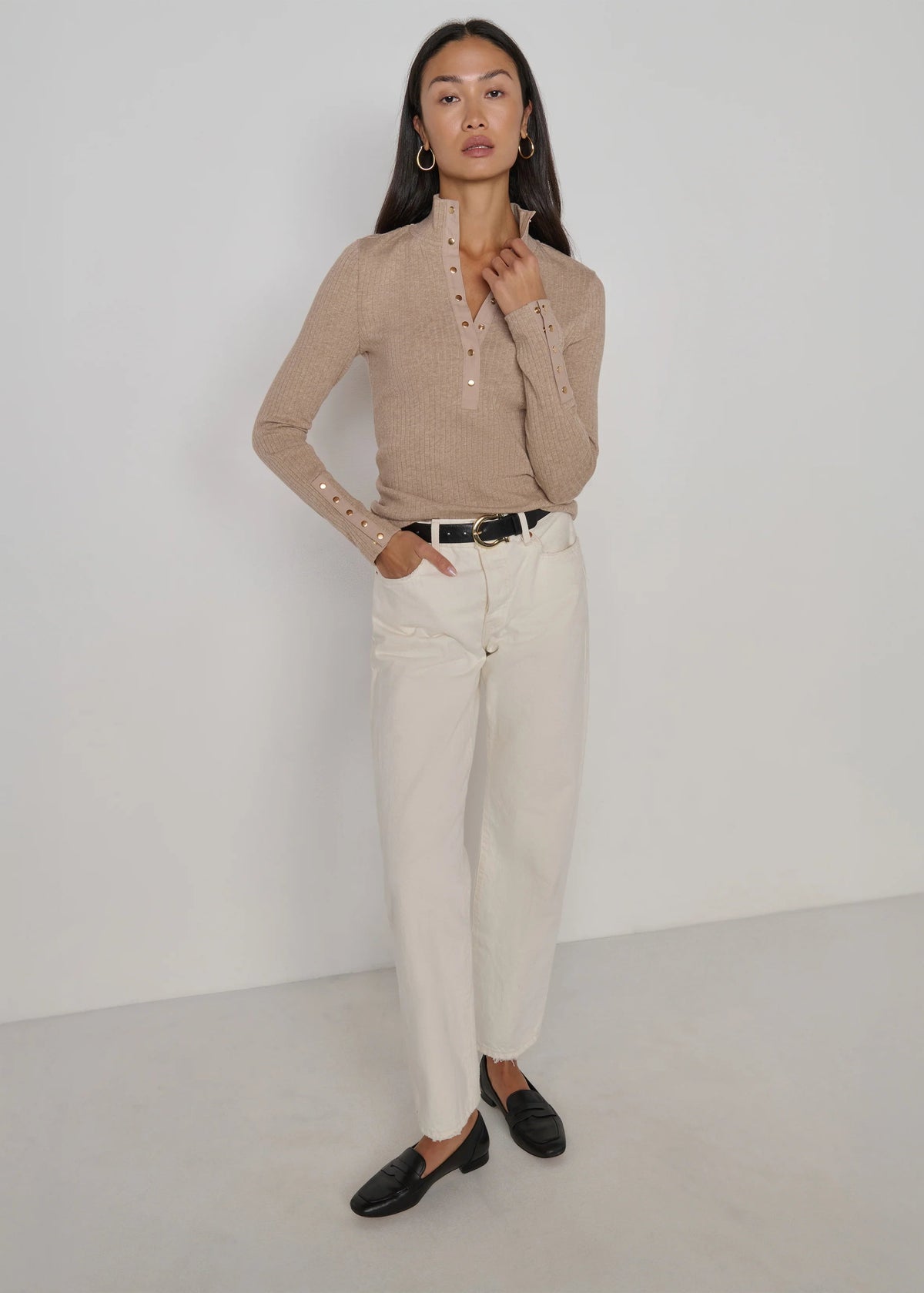 A woman modeling a beige ribbed knit long-sleeve top with decorative buttons on the collar and cuffs, paired with off-white trousers, a black belt, and black loafers, standing against a plain light gray studio background.