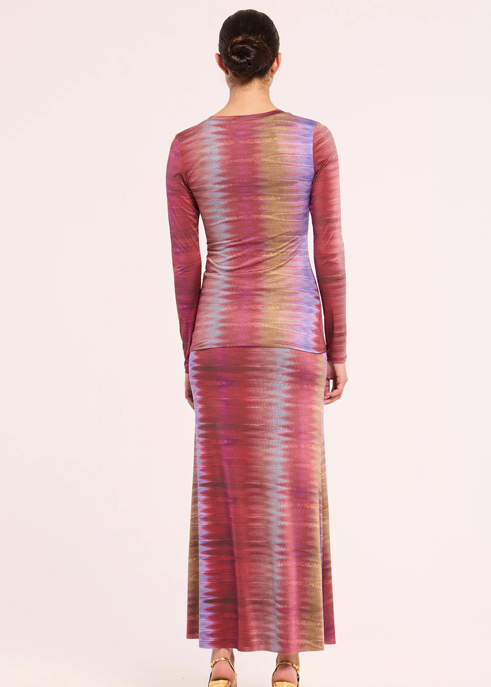 A woman viewed from the back wearing a long-sleeve maxi dress with a vertical tie-dye pattern in pink, purple, blue, and beige tones, set against a plain light background.