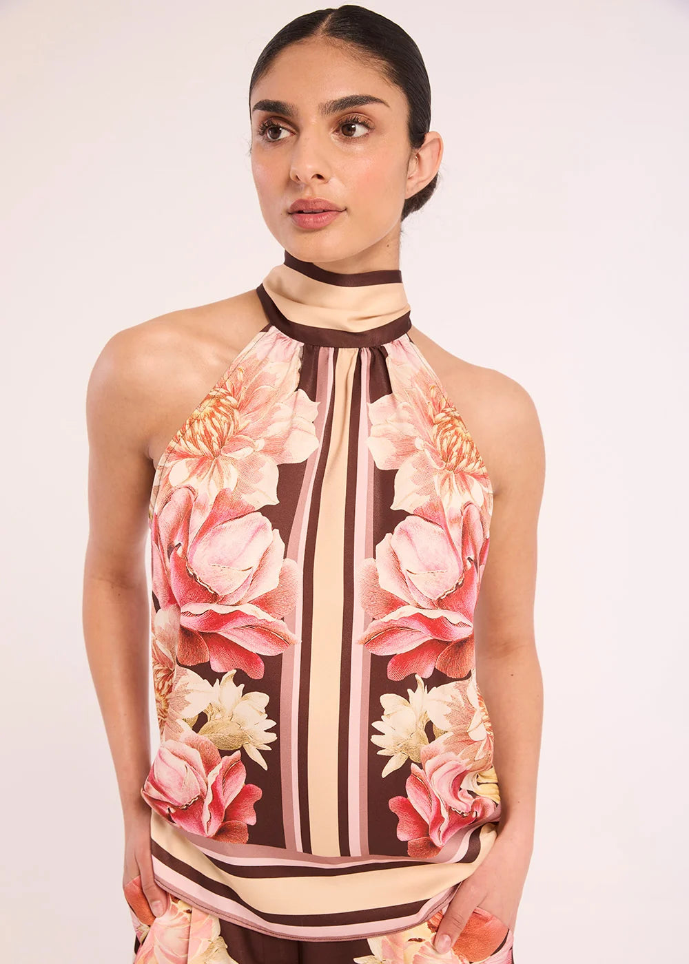 A woman modeling a sleeveless halter top with a brown, beige, and pink floral print, paired with matching floral pants against a plain light background.
