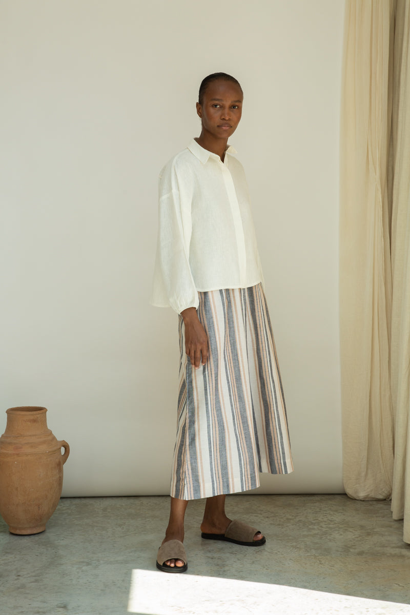 Person wearing a white shirt and striped skirt standing in a minimalistic room with a vase and curtain.