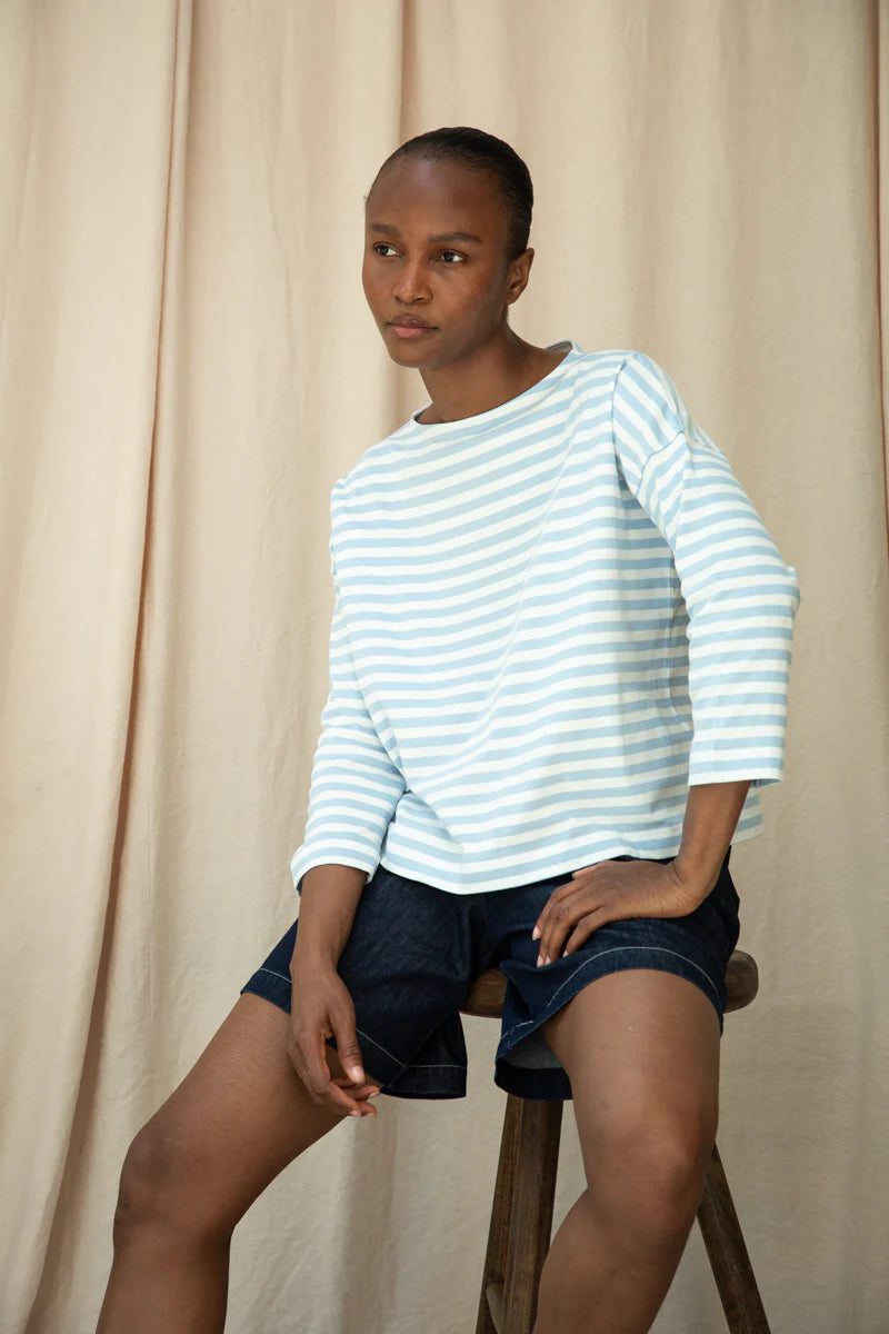 Woman wearing a light blue and white striped shirt and dark shorts sitting on a wooden stool against a beige curtain.