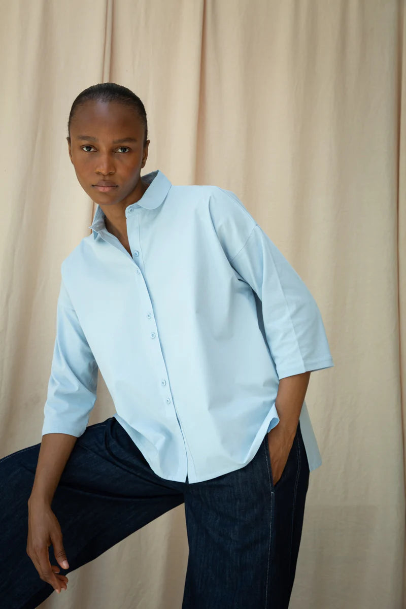 Person wearing a light blue shirt and dark pants against a beige curtain background