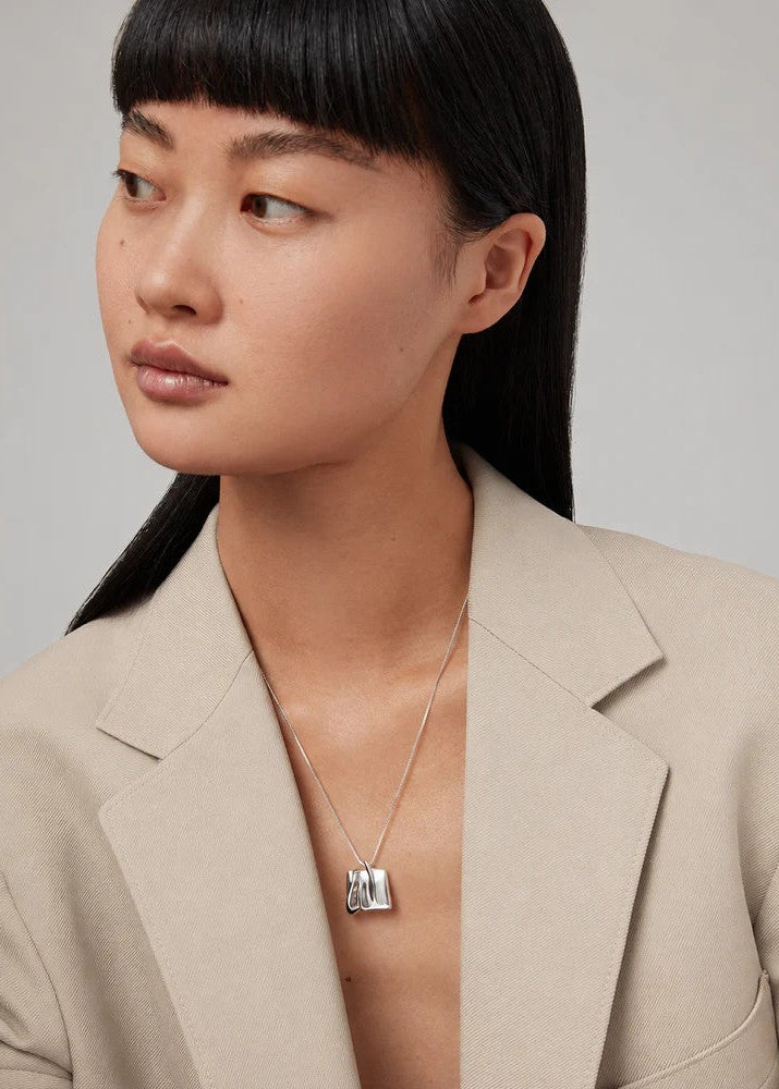 A close-up of a model wearing a silver geometric pendant necklace with a clear stone, styled with a beige blazer.