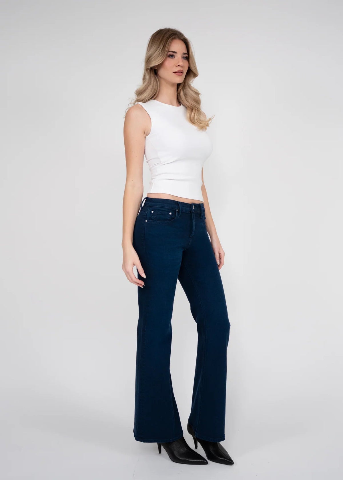 A woman modeling a pair of dark blue flared jeans with a white sleeveless crop top and black heels, standing against a light gray studio background.