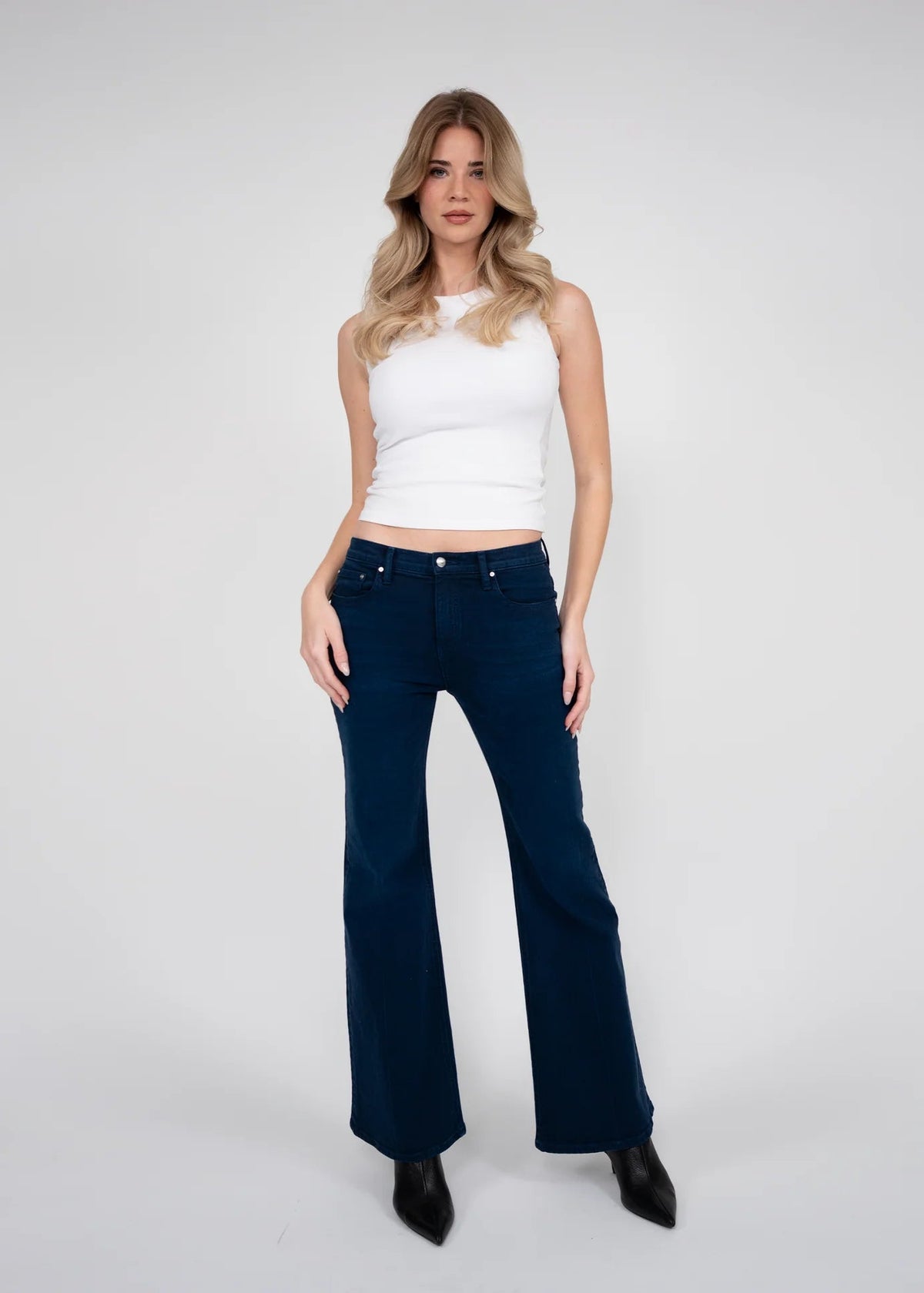 A woman modeling dark navy blue flare jeans with a white ribbed tank top and black pointed-toe heels, standing against a light gray studio background.