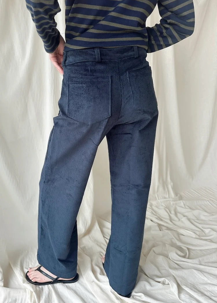A person shown from the back wearing dark blue wide-leg trousers and a navy and beige striped long-sleeve shirt, standing on a white fabric backdrop.