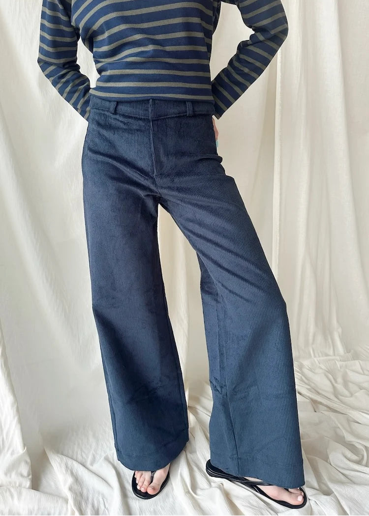 A person wearing dark blue wide-leg corduroy pants and a navy and beige striped long-sleeve top, standing against a draped white fabric backdrop.