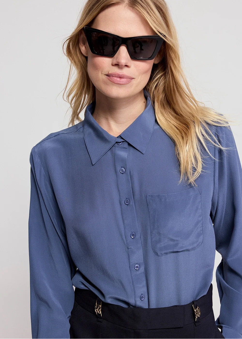 A woman wearing a slate blue long-sleeve button-down shirt and black sunglasses, cropped at the waist with black trousers. The photo is taken against a clean, light gray studio background.