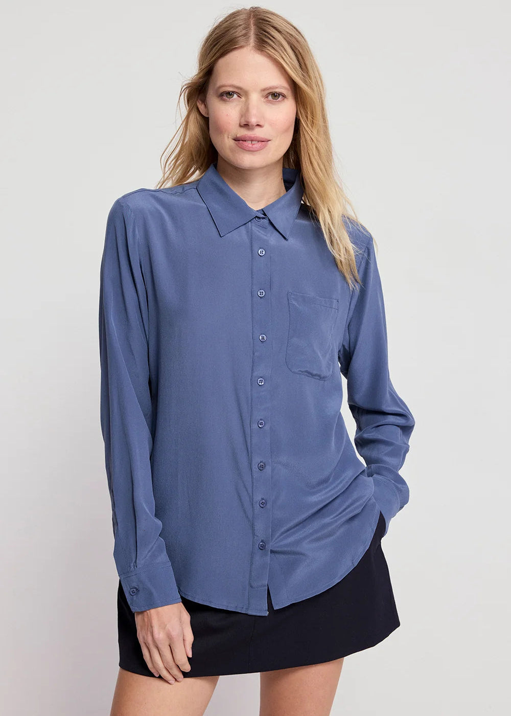 A woman modeling a slate blue long-sleeve button-down shirt with a chest pocket, paired with a black skirt, against a light gray studio background.