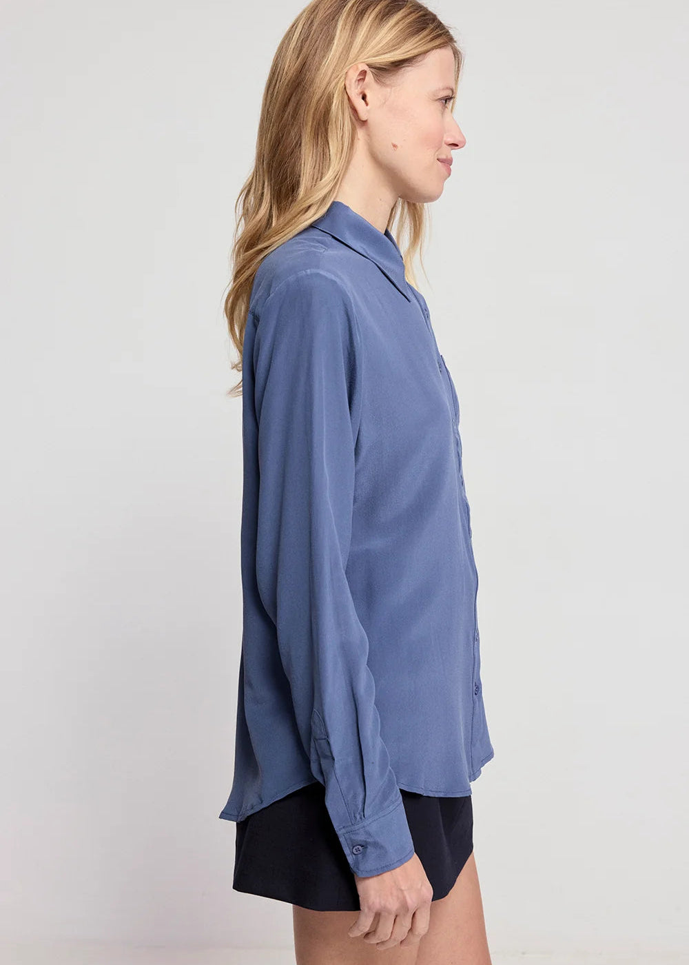 A woman shown in side profile wearing a loose, long-sleeve button-up shirt in dusty medium blue with black shorts, photographed against a plain light gray studio background.