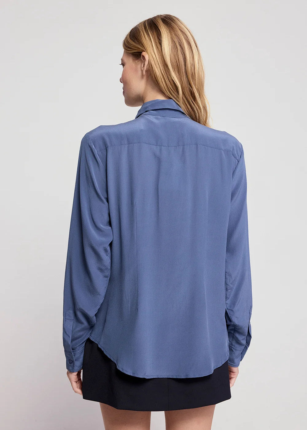 A woman viewed from the back wearing a slate blue button-down shirt and a black skirt, set against a plain light gray studio background.