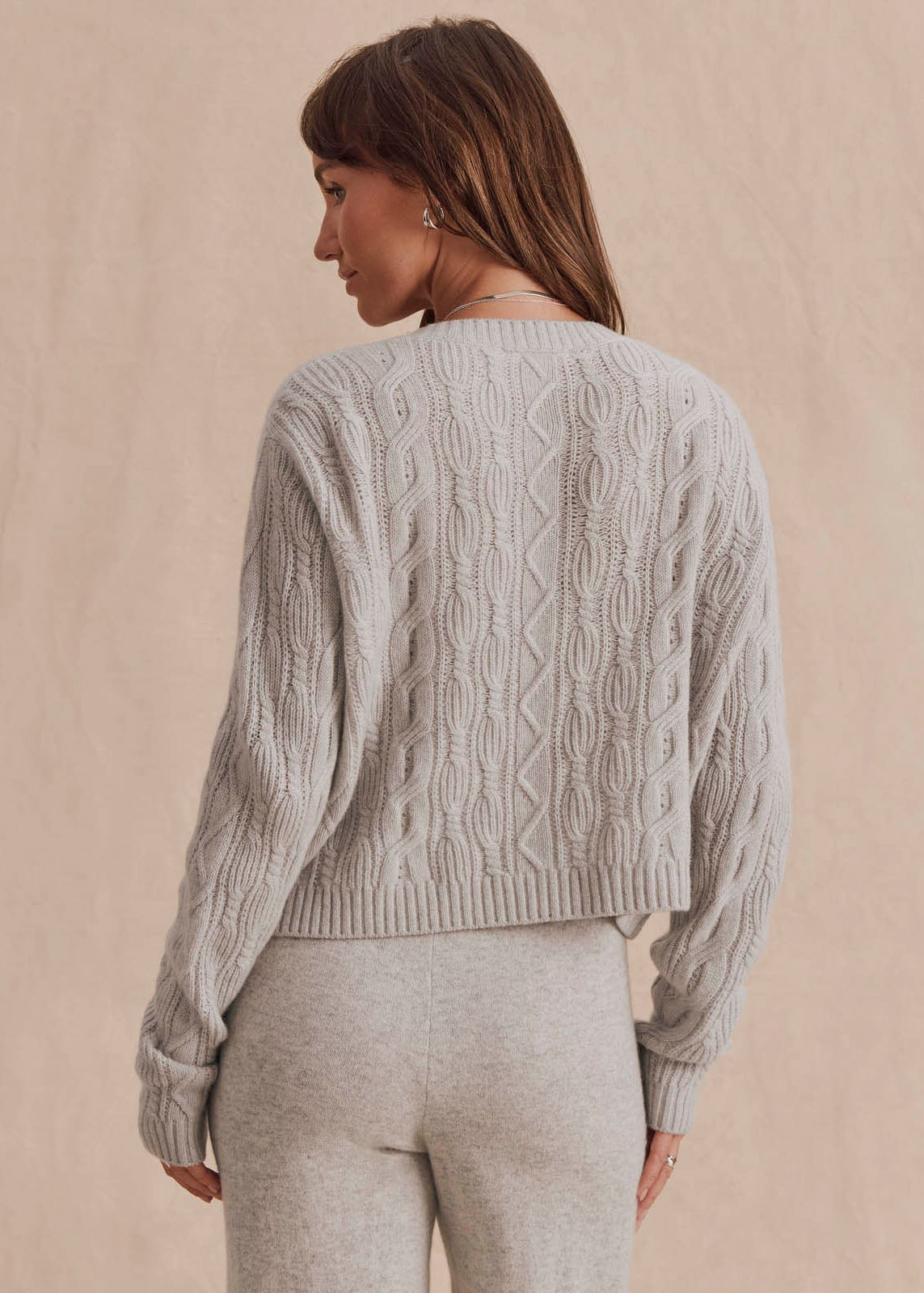A woman shown from the back wearing a light gray cable-knit cropped sweater and matching light gray pants against a soft beige studio background.